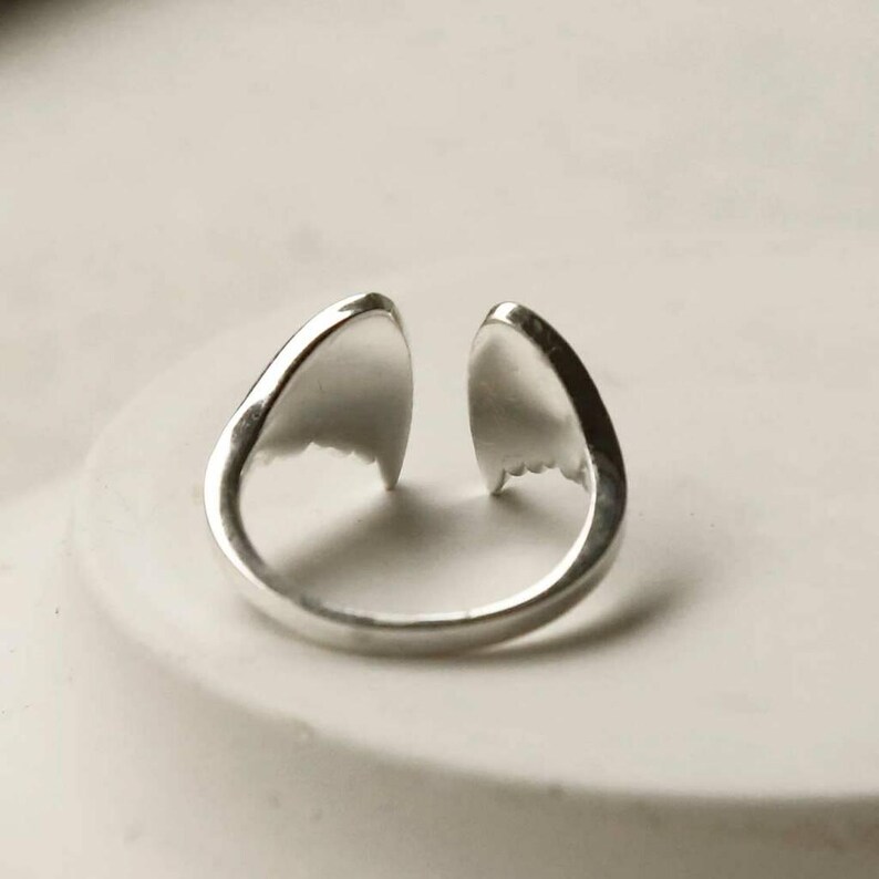 May include: A silver-toned open ring featuring two symmetrical, pointed, wing-like elements that face each other at the top. The band is smooth and polished, creating a continuous loop that remains open at the center, showcasing a metallic finish.