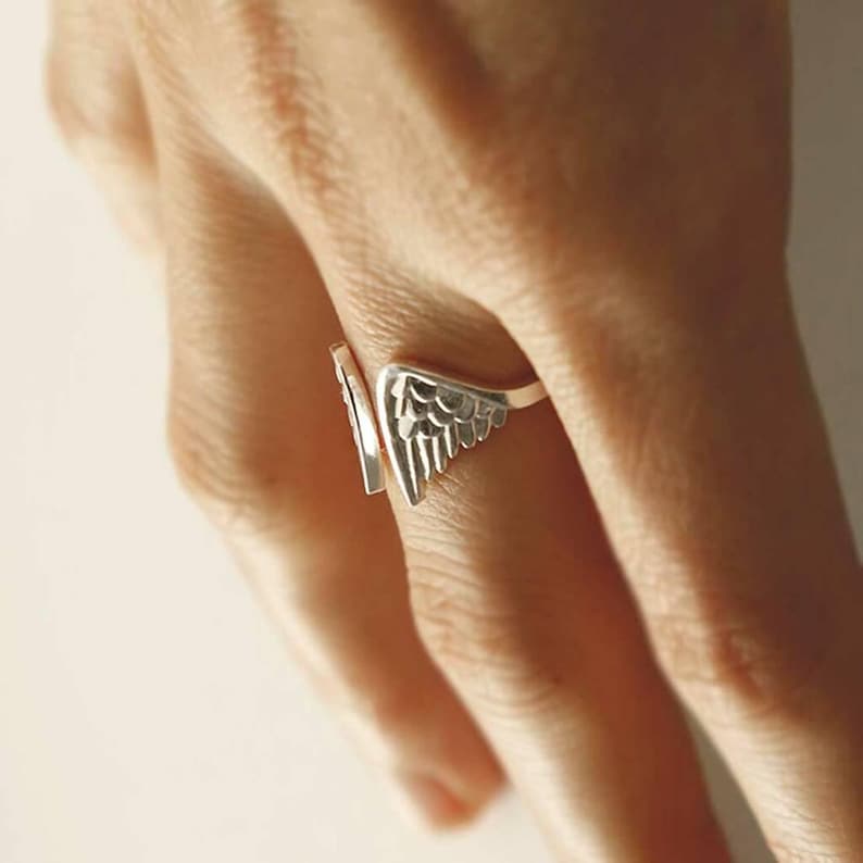 May include: A silver-toned open ring featuring a detailed wing design with layered feather textures. The band is split, allowing the two ends to wrap around the finger, showcasing a polished metallic finish and intricate carved details on the wings.