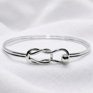 May include: A polished silver-tone bangle bracelet featuring a central infinity-style knot design. The bracelet has a smooth, reflective metallic finish and a circular band that connects to the knot detail, which includes a small spherical accent.