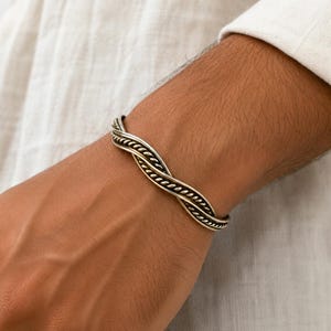 Puede incluir: Una pulsera tipo brazalete de tono plateado con un diseño de cuerda trenzada que se curva en forma de onda. El metal tiene un acabado pulido con ranuras oscurecidas que resaltan la textura intrincada de los hilos trenzados a lo largo de la banda.