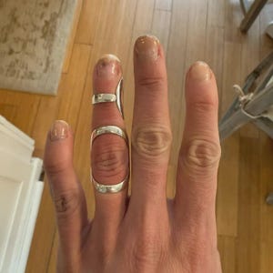 May include: A silver-colored metal finger splint worn on the middle finger. The splint features a smooth, curved design with two open bands that wrap around the finger joint, providing a structured, metallic support for the digit.