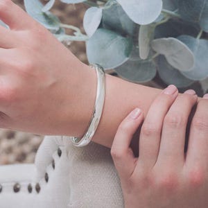 May include: A smooth, polished silver bangle bracelet with a rounded, solid profile. The metal surface is highly reflective, showing a bright metallic sheen. The bangle features a simple, continuous circular design without any additional embellishments.