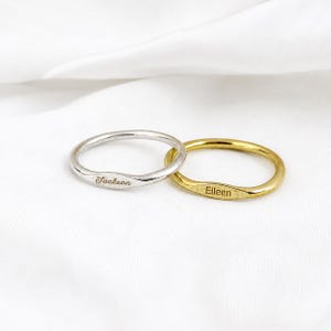 Puede incluir: Dos anillos de metal finos, uno de tono plateado y otro de tono dorado, cada uno con una pequeña sección central plana y ovalada. El anillo plateado está grabado con el nombre Jackson y el dorado con el nombre Eileen en letra cursiva.