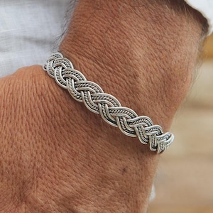 May include: A silver bracelet featuring a braided design. The bracelet is composed of interwoven strands, forming a textured and detailed pattern. The bracelet is worn on a wrist.