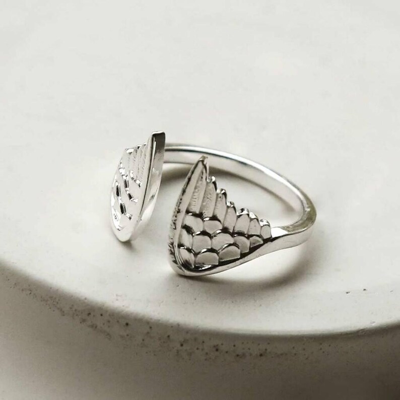 May include: A silver-toned open ring featuring two symmetrical wing-shaped ends. Each wing element is detailed with embossed, layered feather patterns that curve inward toward the center gap of the band, creating a polished metallic finish.
