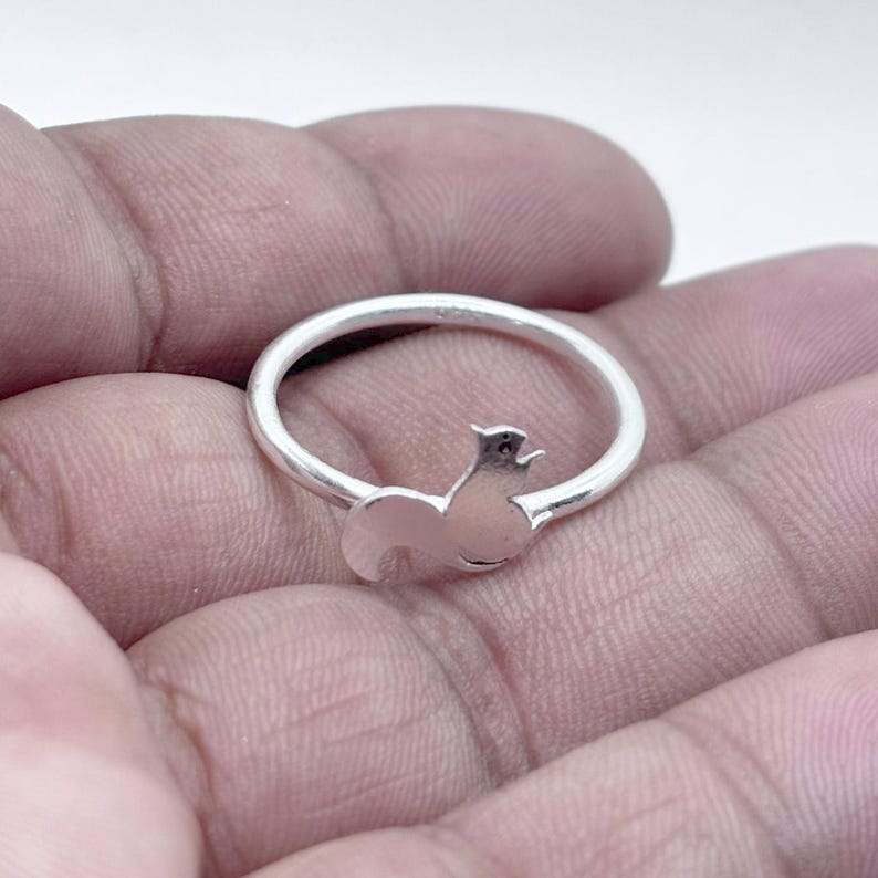 May include: A polished silver ring featuring a small, stylized squirrel silhouette integrated into the band. The metal has a smooth, reflective finish, and the design shows the squirrel in a seated position with its tail curved upward along the ring.