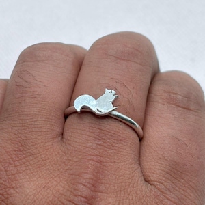 May include: A silver-toned ring featuring a small, flat, squirrel-shaped charm attached to the top of the thin, circular band. The squirrel silhouette is oriented horizontally, showcasing a curved tail and a small head, with a smooth, metallic finish.