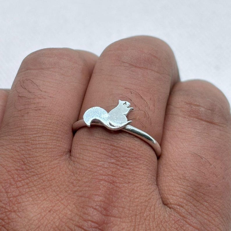 May include: A silver-toned ring featuring a small, flat, squirrel-shaped charm attached to the top of the thin, circular band. The squirrel silhouette is oriented horizontally, showcasing a curved tail and a small head, with a smooth, metallic finish.