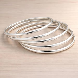 May include: A set of five smooth, polished silver-toned bangle bracelets stacked together. Each bracelet features a simple, rounded, continuous circular band design with a reflective metallic finish that catches the light, creating a uniform appearance.