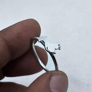May include: A silver-toned ring featuring a small, flat, cutout silhouette of a squirrel perched on the top of the band. The squirrel design is oriented horizontally, showing the animal's body, head, and curved tail in a simple, polished metal finish.