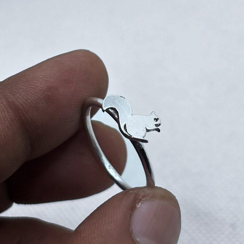 May include: A silver-toned ring featuring a small, flat, cutout silhouette of a squirrel perched on the top of the band. The squirrel design is oriented horizontally, showing the animal's body, head, and curved tail in a simple, polished metal finish.