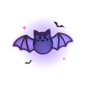 May include: A purple bat-shaped cookie cutter with a black outline. The central body features a small, simple face with two circular eyes and a curved mouth. Two small black bat silhouettes and two pink four-pointed stars surround the main shape.