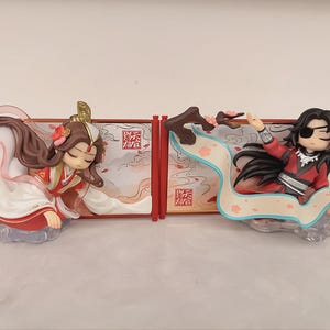 May include: Two chibi-style anime figurines featuring characters in traditional red and white robes. One figure has long brown hair and a gold headpiece, while the other has black hair, an eyepatch, and a flowing ribbon. Both stand before red-framed panels.