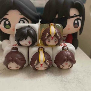 Puede incluir: Una colección de cinco dados cuadrados con rostros de personajes estilizados, cabello oscuro, tocados intrincados y rasgos expresivos. Dos figuras estilo chibi más grandes con cabello negro y rostros detallados se encuentran detrás de los dados.