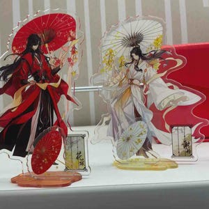 May include: Two acrylic standees featuring anime-style characters in traditional robes. One character wears red with a red umbrella, the other wears white with a yellow umbrella. Each standee includes a small base and a rectangular nameplate.