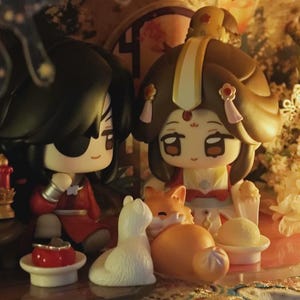May include: Two stylised chibi figurines with dark hair and traditional-style clothing, one wearing a red outfit and the other a brown and white ensemble. They are positioned near a small white cat figurine and a small orange fox figurine.