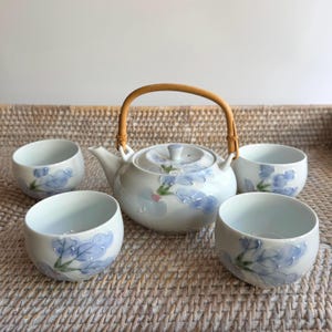 May include: A white ceramic teapot with a curved bamboo handle and four matching white ceramic teacups. Each piece features a hand-painted design of light blue flowers with green stems and small pink accents on the petals and leaves.