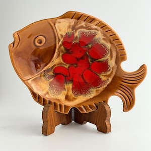 May include: A fish-shaped ceramic dish featuring a glossy brown glaze on the head and tail, with a vibrant red and cream-colored mottled glaze pattern on the body. The dish is displayed on a small, dark brown wooden stand with three legs.
