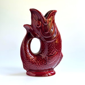 May include: A deep red ceramic pitcher shaped like a fish with an open mouth at the top. The body features a textured scale pattern, a curved tail forming a handle, and a circular opening in the center. The glossy finish reflects light.