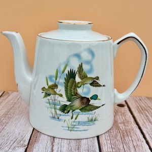 May include: White ceramic teapot with a lid, featuring a painted scene of ducks in flight over water and reeds. The teapot has a gold trim around the rim and handle. The spout and handle are white.