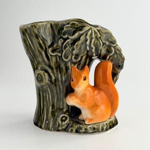 Può includere: Un vaso in ceramica a forma di tronco d'albero verde scuro con corteccia testurizzata e foglie di quercia, caratterizzato da una statuetta di scoiattolo arancione brillante applicata sul davanti. Lo scoiattolo è seduto e tiene una noce.