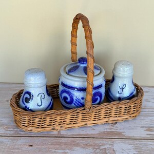 May include: A ceramic salt and pepper set and a lidded jar with a blue and white design, nestled in a woven wicker basket with a handle. The salt and pepper shakers are labeled with the letters "S" and "P".