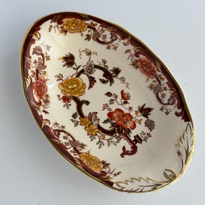 May include: An oval ceramic dish featuring a cream-coloured base with a decorative border in deep burgundy and gold. The centre is adorned with intricate floral patterns in shades of orange, yellow, and brown, accented with gold leaf detailing.