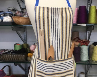 May include: A white and blue striped apron with a large pocket on the front. The apron has a blue strap and is being displayed on a mannequin. The apron is surrounded by spools of yarn in various colours.
