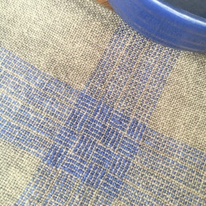 May include: A close-up of a woven linen kitchen towel with a blue and grey geometric pattern. The towel is folded in half and has a blue bowl on top of it.