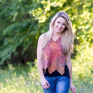 May include: A woman wearing a sleeveless, multi-colored woven top with a black tank top underneath and blue jeans. She is standing in a field of green grass.