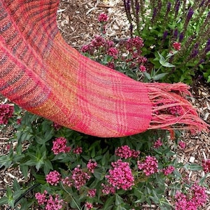 Puede incluir: Un chal tejido rojo y naranja con flecos, drapeado sobre un lecho de flores rosas.