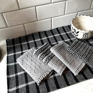 May include: Four grey and black woven kitchen towels with fringed edges on a black and white checked placemat. A white bowl with black floral design is on the placemat.