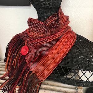 Puede incluir: Una bufanda tejida en tonos naranja, rojo y marrón con un botón rojo. La bufanda tiene flecos en los extremos.