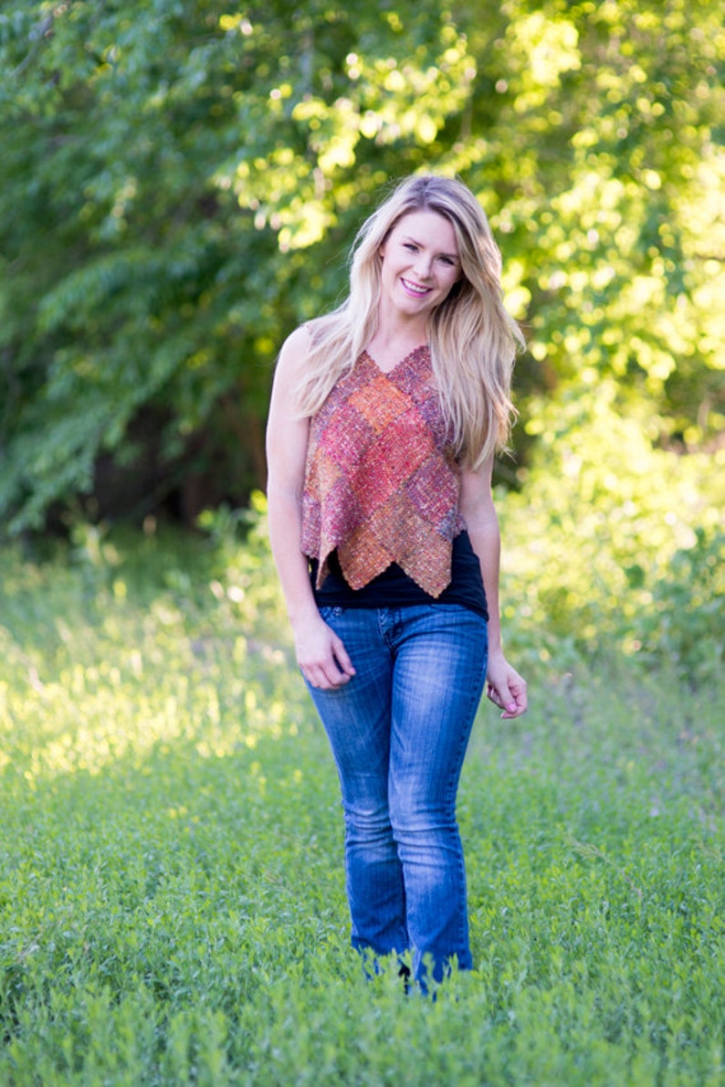 May include: A woman wearing a sleeveless, multi-colored woven top with a black tank top underneath and blue jeans. She is standing in a field of green grass.