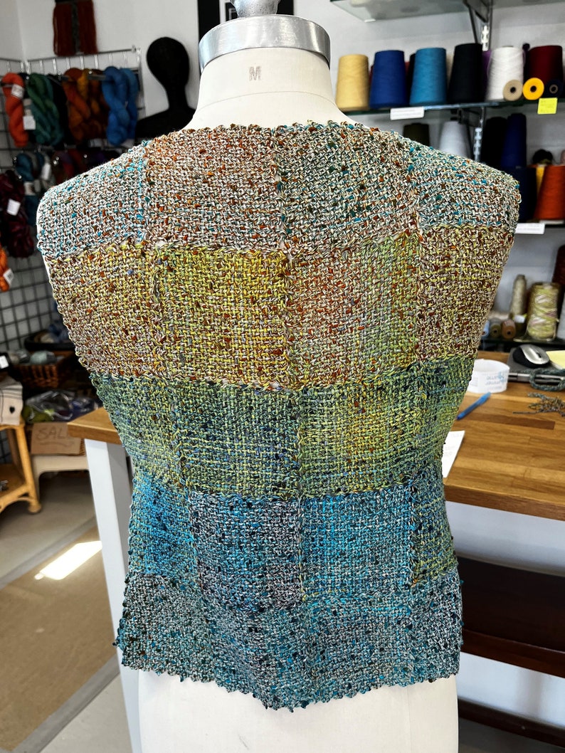 May include: A woven vest with a multi-colored pattern of blue, green, yellow, and brown. The vest is made of a textured fabric and has a slightly frayed edge.