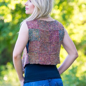 May include: A woman wearing a sleeveless vest with a multi-colored woven pattern. The vest is cropped and has a relaxed fit. The woman is wearing blue jeans and a black tank top.