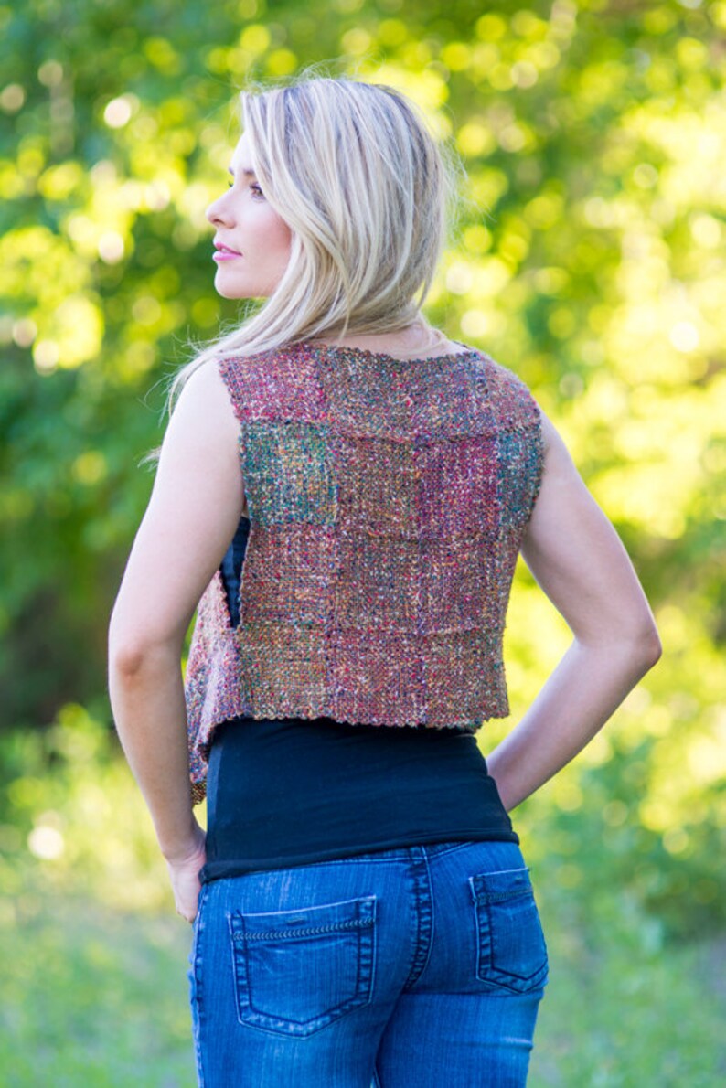 May include: A woman wearing a sleeveless vest with a multi-colored woven pattern. The vest is cropped and has a relaxed fit. The woman is wearing blue jeans and a black tank top.