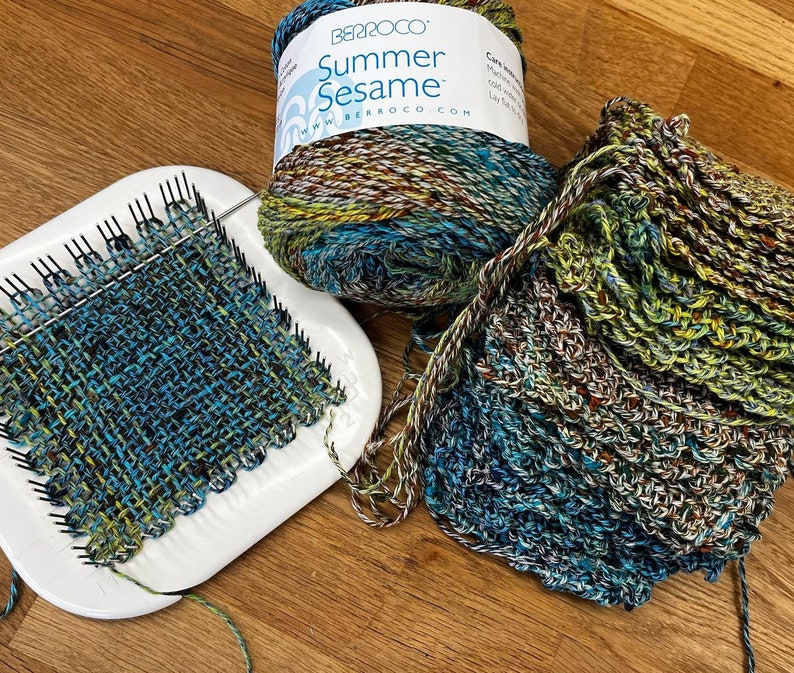 May include: A close-up of a loom with a woven fabric in shades of blue, green, and yellow. The yarn is in a skein with the label "Summer Sesame" and the brand name "Berroco".