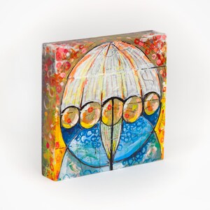 May include: A rectangular wooden block featuring a painted illustration of a white umbrella with yellow circular accents over a blue textured base. The background displays vibrant orange, red, and yellow abstract patterns with visible brushstrokes.