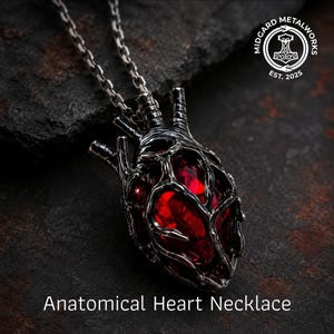 May include: An anatomical heart necklace featuring a dark, oxidised silver-toned metal cage surrounding a faceted, deep red gemstone. The pendant hangs from a matching silver-toned chain. The text "Midgard Metalworks Est. 2025" and "Anatomical Heart Necklace" are visible.