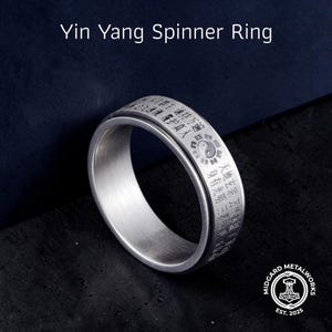 May include: A silver-colored Yin Yang spinner ring. The ring features engraved Chinese characters and a Yin Yang symbol. The text "Yin Yang Spinner Ring" is at the top. The ring is on a dark blue background.