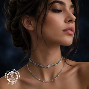 May include: A layered silver-toned necklace set featuring a flat curb chain choker with a central round white stone and a secondary delicate cable chain adorned with small, evenly spaced clear beads. A circular logo with a hammer icon reads MIDGARD METALWORKS EST. 2025.