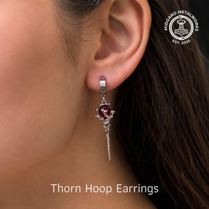 May include: A silver-toned hoop earring featuring a dangling charm with a dark red spherical bead encased in a metallic, thorn-like structure. The earring hangs from a simple hoop closure. The text Midgard Metalworks Est. 2025 is visible.