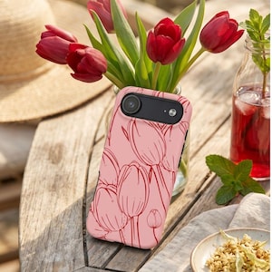 May include: A pink phone case featuring a red tulip design sits on a wooden table. A vase of red tulips and a straw hat are in the background. A glass bottle with a red liquid and a small plate with dried herbs are also visible.