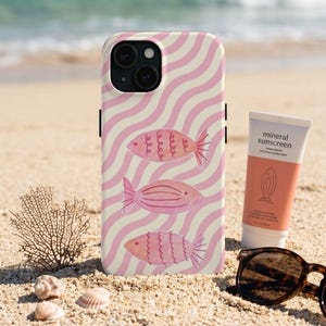Könnte beinhalten: Ein Handyhülle mit einem rosa-weißen Wellenmuster und drei rosa Fischillustrationen. Eine Tube mineralische Sonnencreme und eine Schildpatt-Sonnenbrille liegen auf dem Sand. Auch Korallen und Muscheln sind am Strand zu sehen.