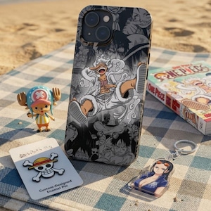 Puede incluir: Una funda para iPhone con un gráfico en blanco y negro de un personaje de la serie de anime One Piece. También se muestra una figura de Chopper, un pin de esmalte, un llavero y un volumen de manga de One Piece.