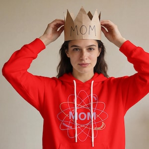 May include: A red hoodie with a white drawstring and a graphic that says "MOM" in the center. The graphic is surrounded by a design with the words "PATIENCE", "STRENGTH", and "CARE". A brown paper crown with the word "MOM" is being held above the head.