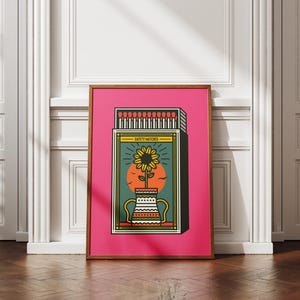May include: Framed art print with a sunflower illustration inside a matchbox. The artwork has a pink background and a wooden frame. The text "SAFETY MATCHES" is visible. The print is displayed on a wooden floor.