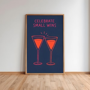 May include: A framed print with the words "CELEBRATE SMALL WINS" in pink above two cocktail glasses filled with orange liquid. The glasses are outlined in pink on a dark blue background. The frame is made of wood.
