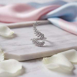 Peut inclure: Une bague argentée ornée d'un motif de feuilles serties de petits brillants transparents. La bague ouverte présente un design de feuilles qui s'enroule autour du doigt. Posée sur une surface en marbre blanc, entourée de pétales de fleurs blanches.